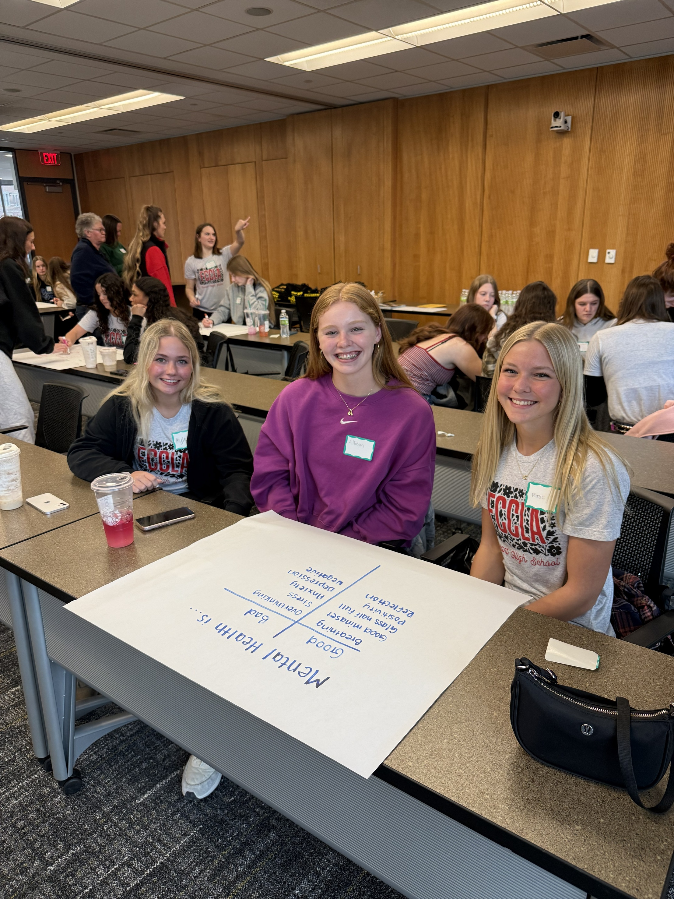 3 teens smiling at camera, poster that states "mental health is" on table in front of them