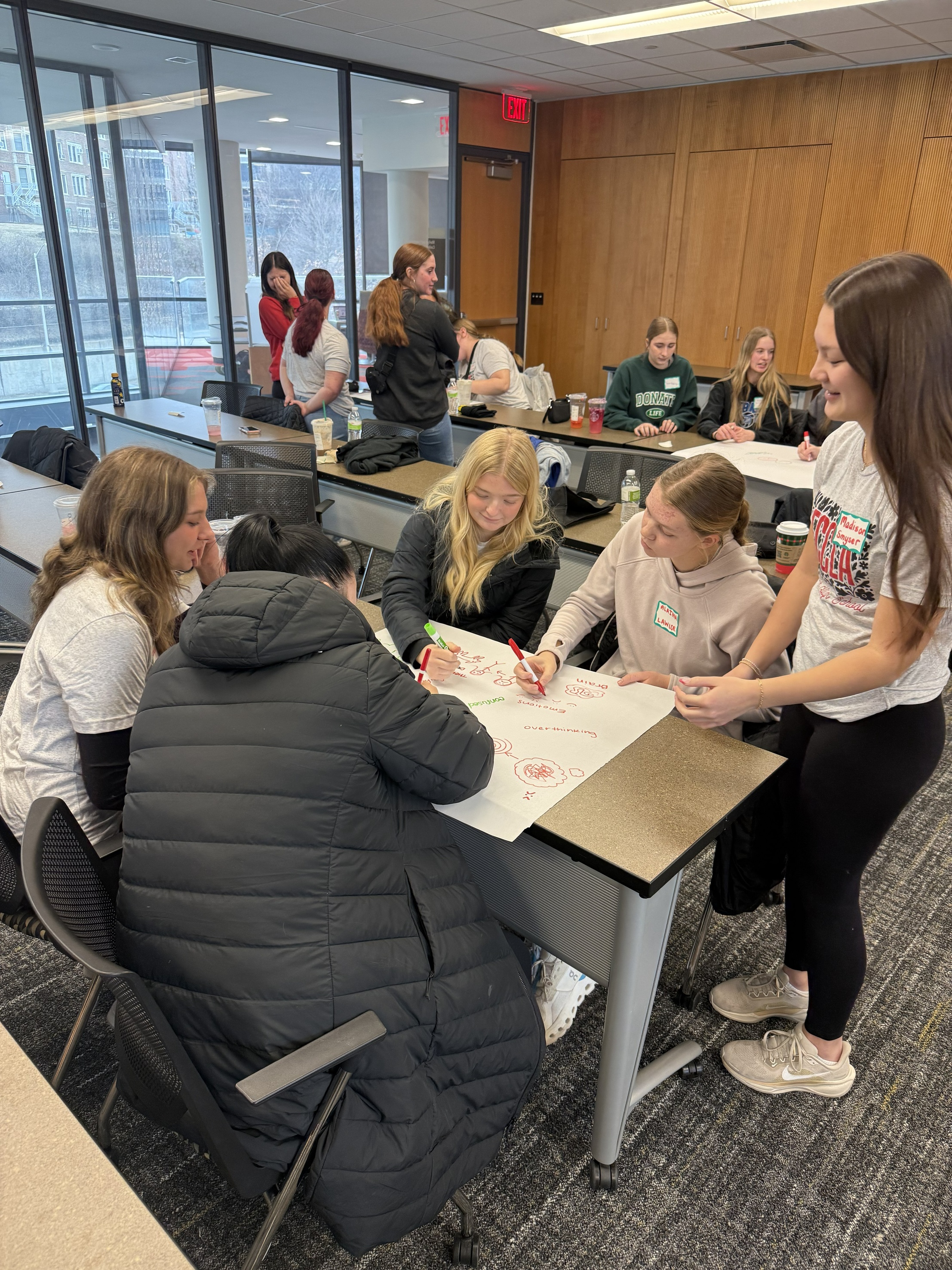 Teens huddled around a table creating a mental health poster, teens lingering in background of photo