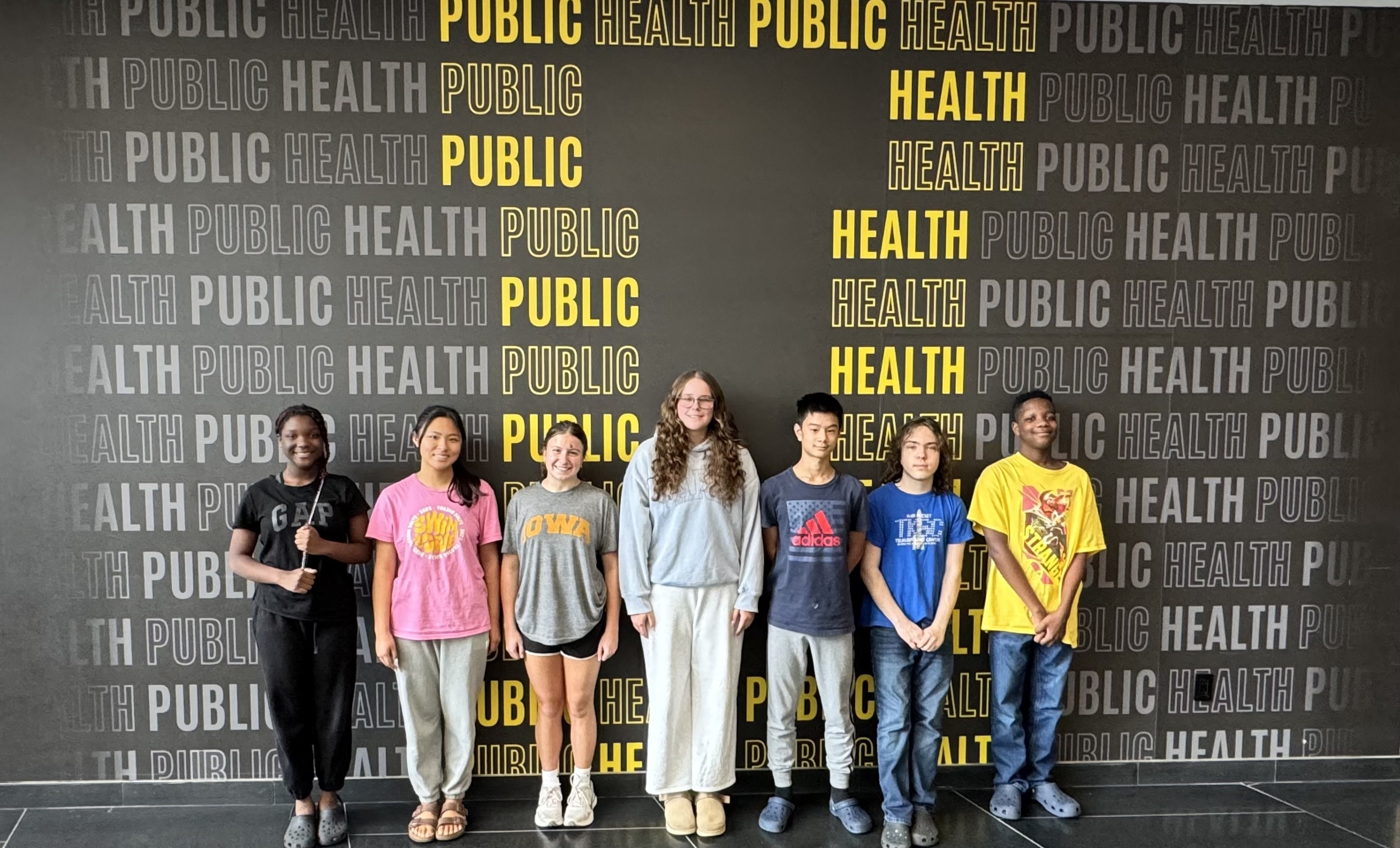 Group of seven youth standing in front of a black wall featuring repeated “PUBLIC HEALTH” text in gray and yellow. The group faces the camera in casual clothing, with the large public health graphic wall serving as the background.